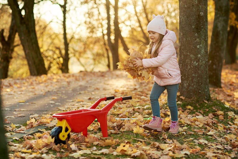 A young girl enjoys piling autumn leaves into a toy wheelbarrow in a sunlit park.
